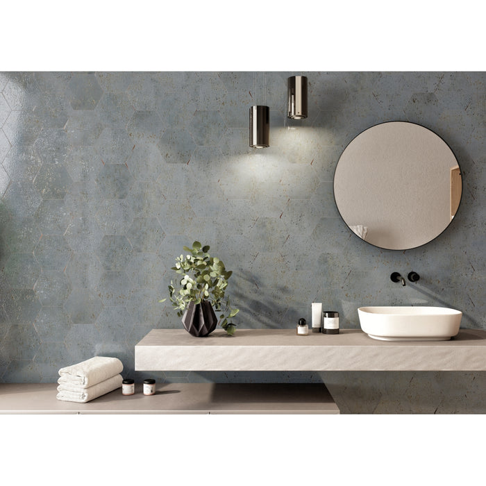 A modern bathroom vanity setup featuring a floating light beige countertop with a white vessel sink, a round wall mirror, and a textured slate-blue wall with two cylindrical wall lights. Decorative greenery and folded towels are arranged on the counter.