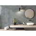 A modern bathroom vanity setup featuring a floating light beige countertop with a white vessel sink, a round wall mirror, and a textured slate-blue wall with two cylindrical wall lights. Decorative greenery and folded towels are arranged on the counter.
